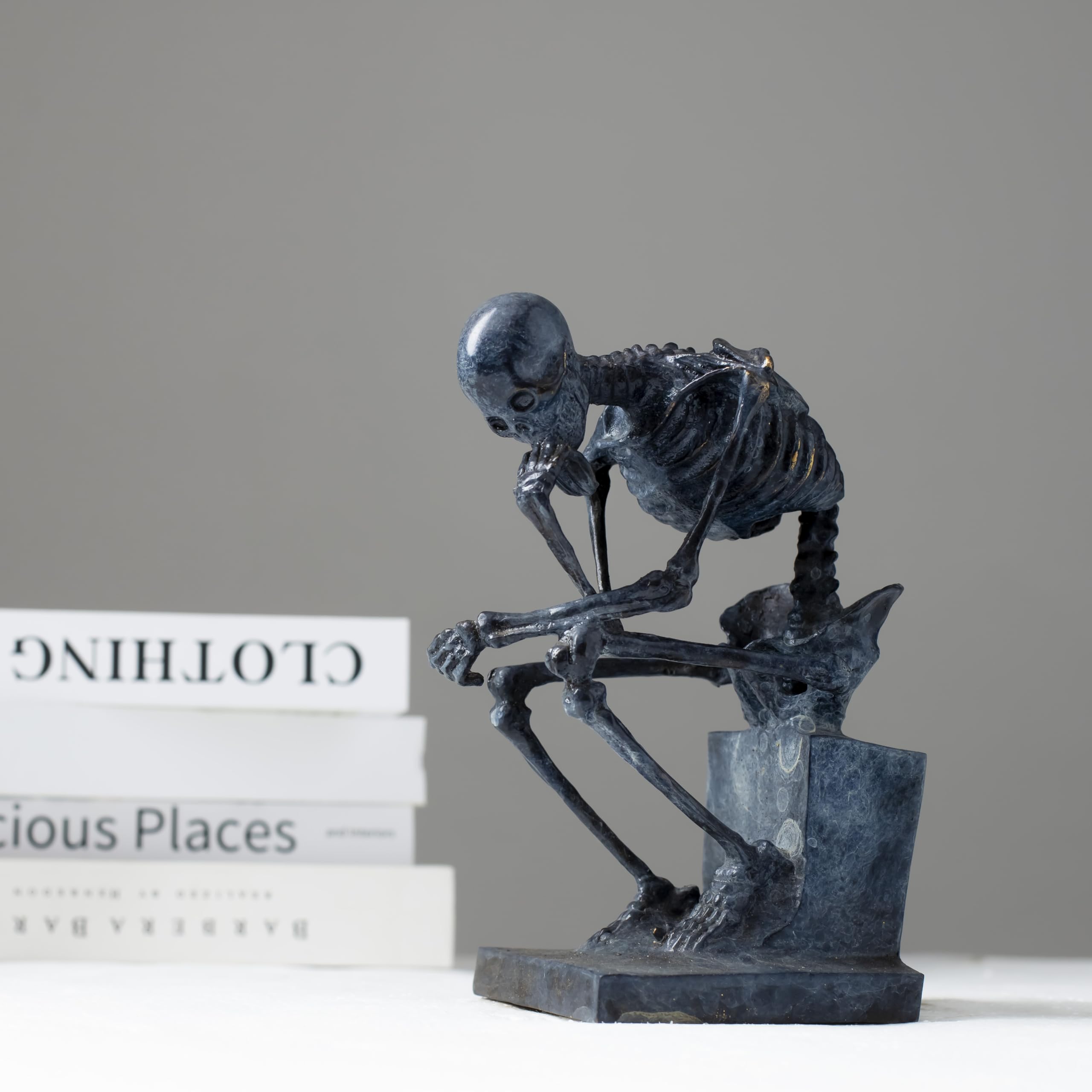 Bronze skeleton Thinker statue in blue-grey patina displayed on bookshelf beside books