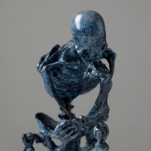 Bronze skeleton Thinker statue in blue-grey patina on bookshelf showing front detail and base