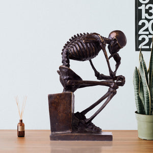 Bronze skeleton Thinker statue in brown bronze finish beside books on light gray background