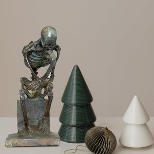 Bronze skeleton Thinker statue in brown bronze finish beside decorative tree ornaments