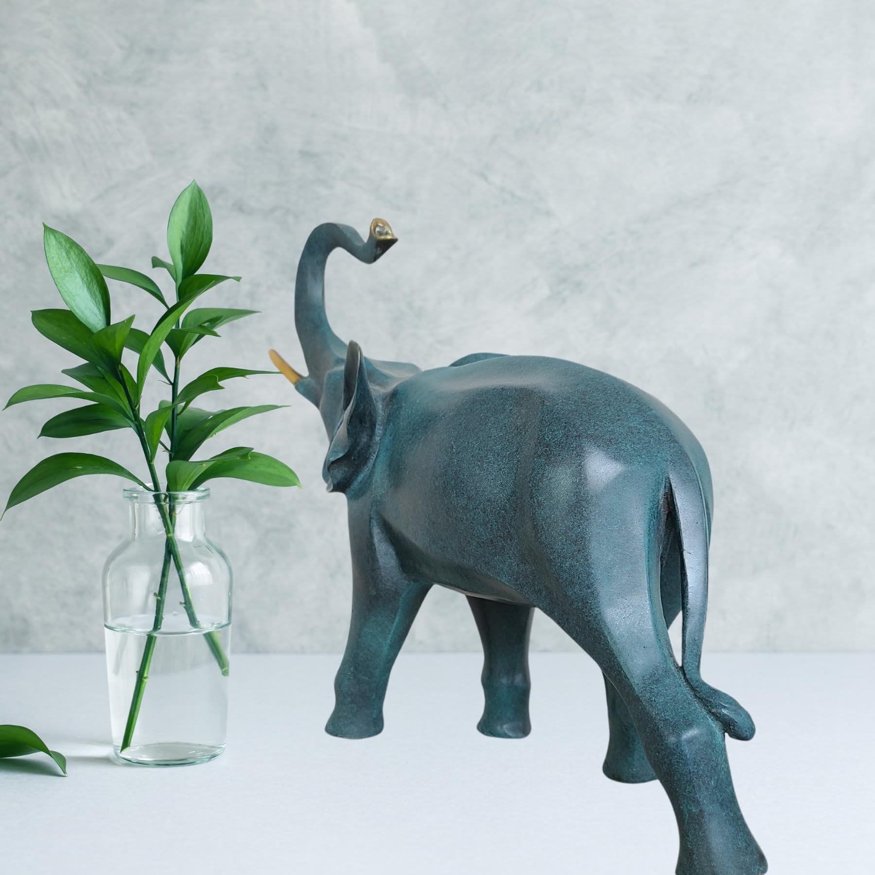 Close-up of blue bronze elephant statue beside green plant against textured gray wall