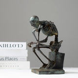 Bronze skeleton Thinker statue in antique grey bronze finish beside books on white surface