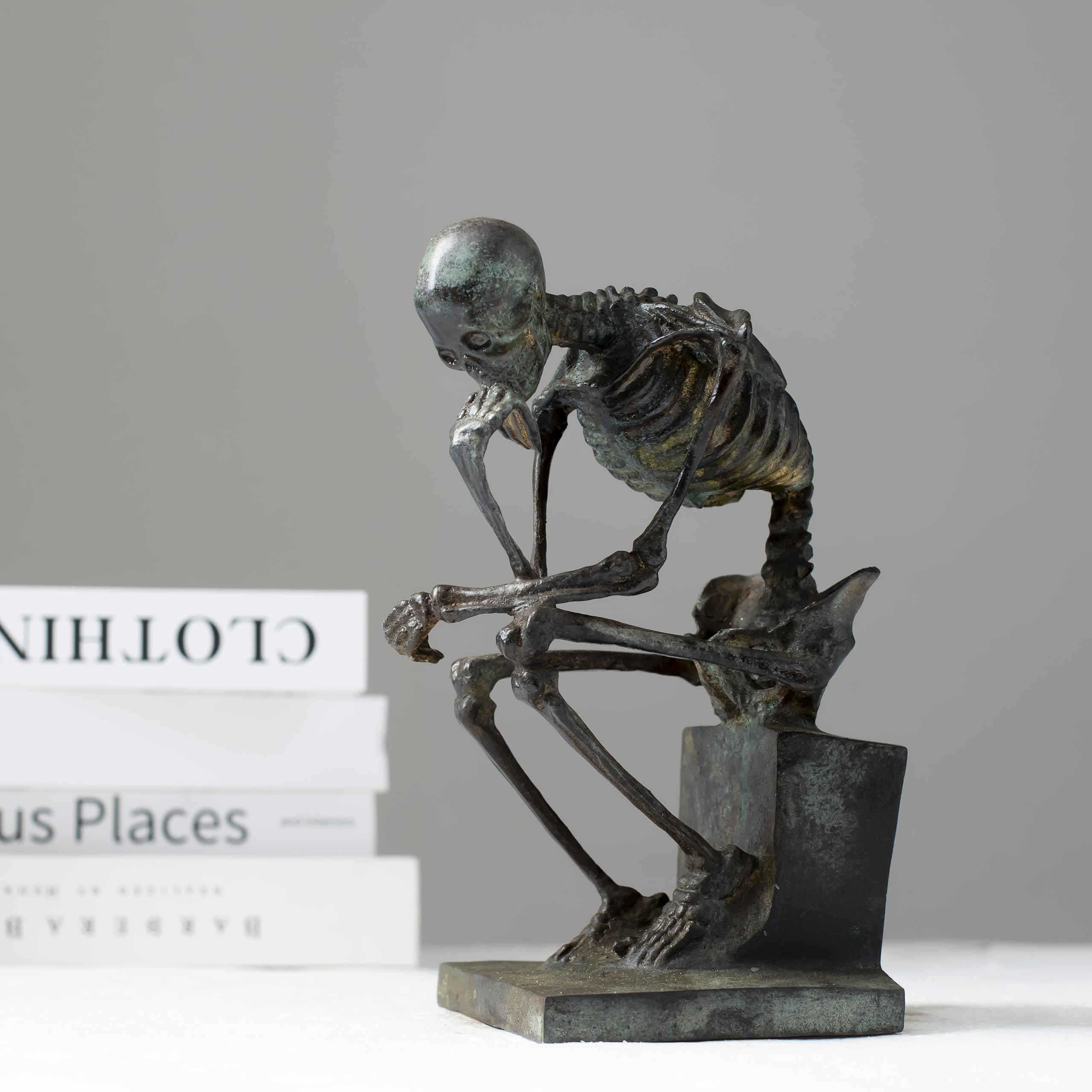 Bronze skeleton Thinker statue in antique grey bronze finish beside books on white surface