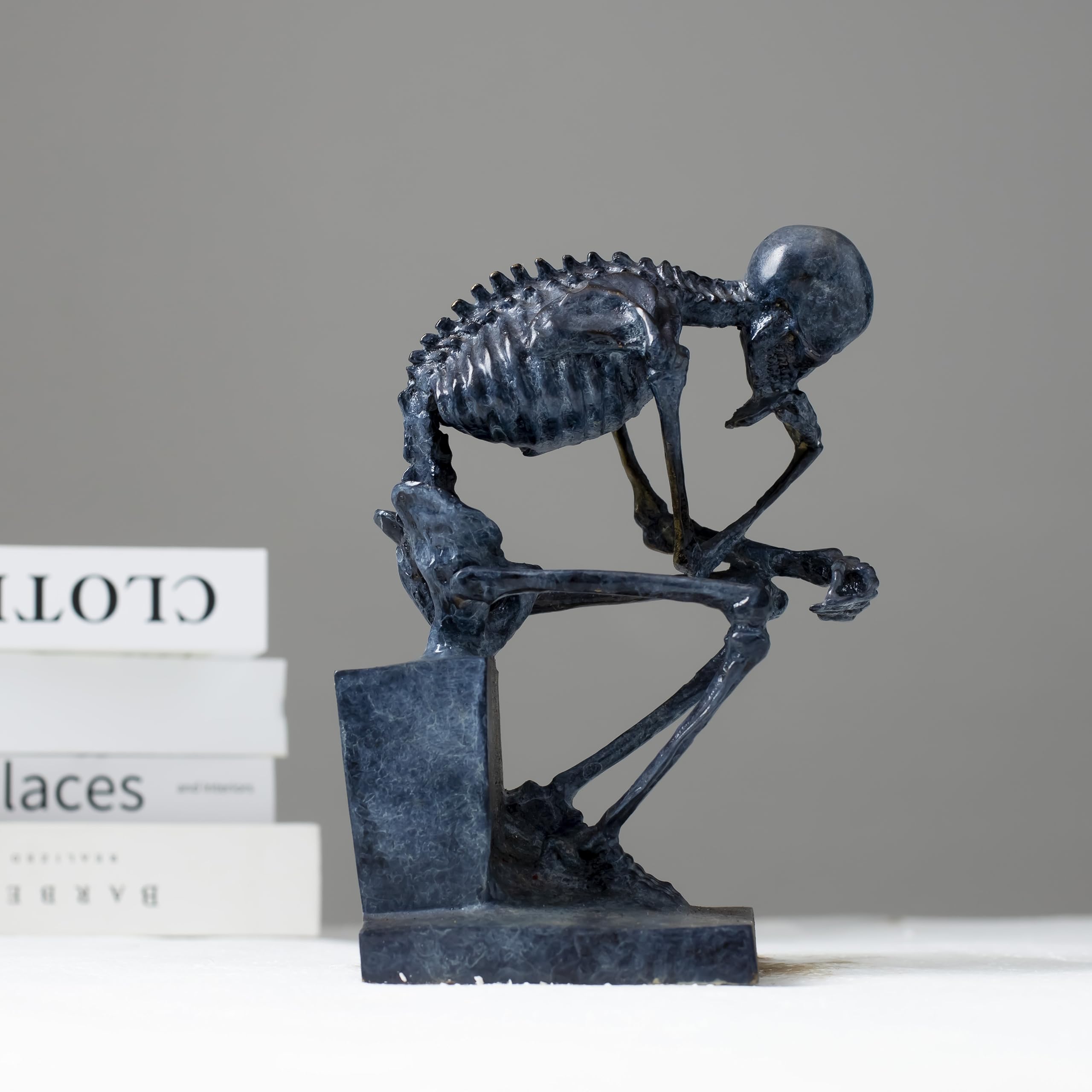 Bronze skeleton Thinker statue in blue-grey patina displayed beside books on white surface