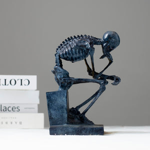 Bronze skeleton Thinker statue in blue-grey patina displayed beside books on white surface