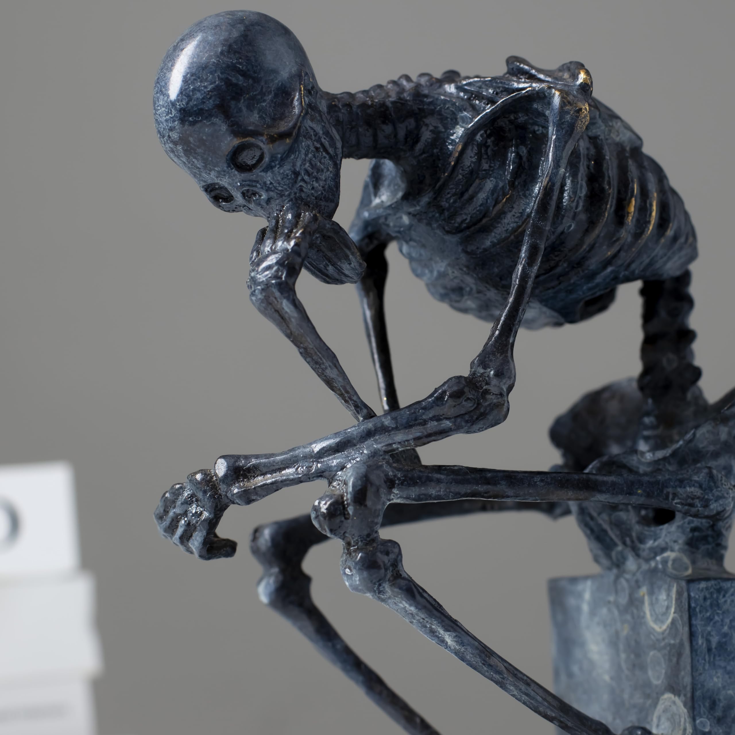 Bronze skeleton Thinker statue in blue-grey patina on bookshelf showing front detail and base