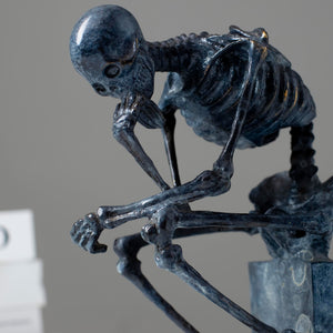 Bronze skeleton Thinker statue in blue-grey patina on bookshelf showing front detail and base