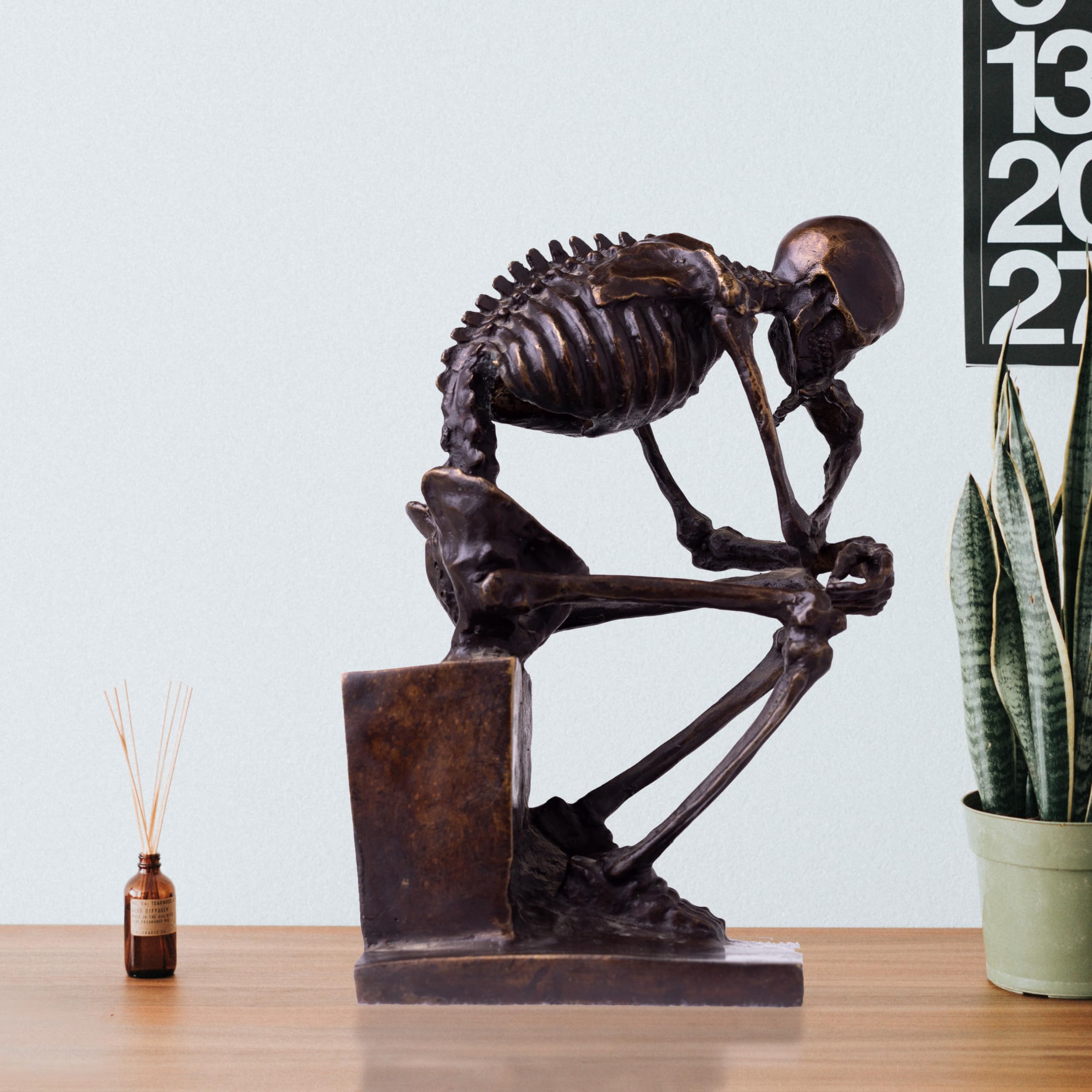 Bronze skeleton Thinker statue in brown bronze finish beside books on light gray background