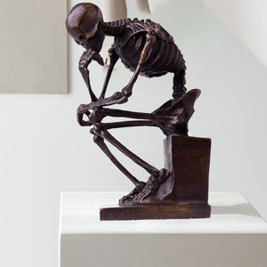 Bronze skeleton Thinker statue in brown bronze finish beside decorative tree ornament