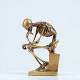 Bronze skeleton Thinker statue in gold finish front view, Rodin-inspired memento mori sculpture on white background