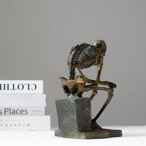 Bronze skeleton Thinker statue in green patina finish displayed on desk beside laptop
