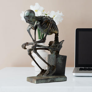 Bronze skeleton Thinker statue in antique grey finish beside decorative glass vase
