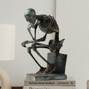 Bronze skeleton Thinker statue displayed on white pedestal showing full seated figure profile