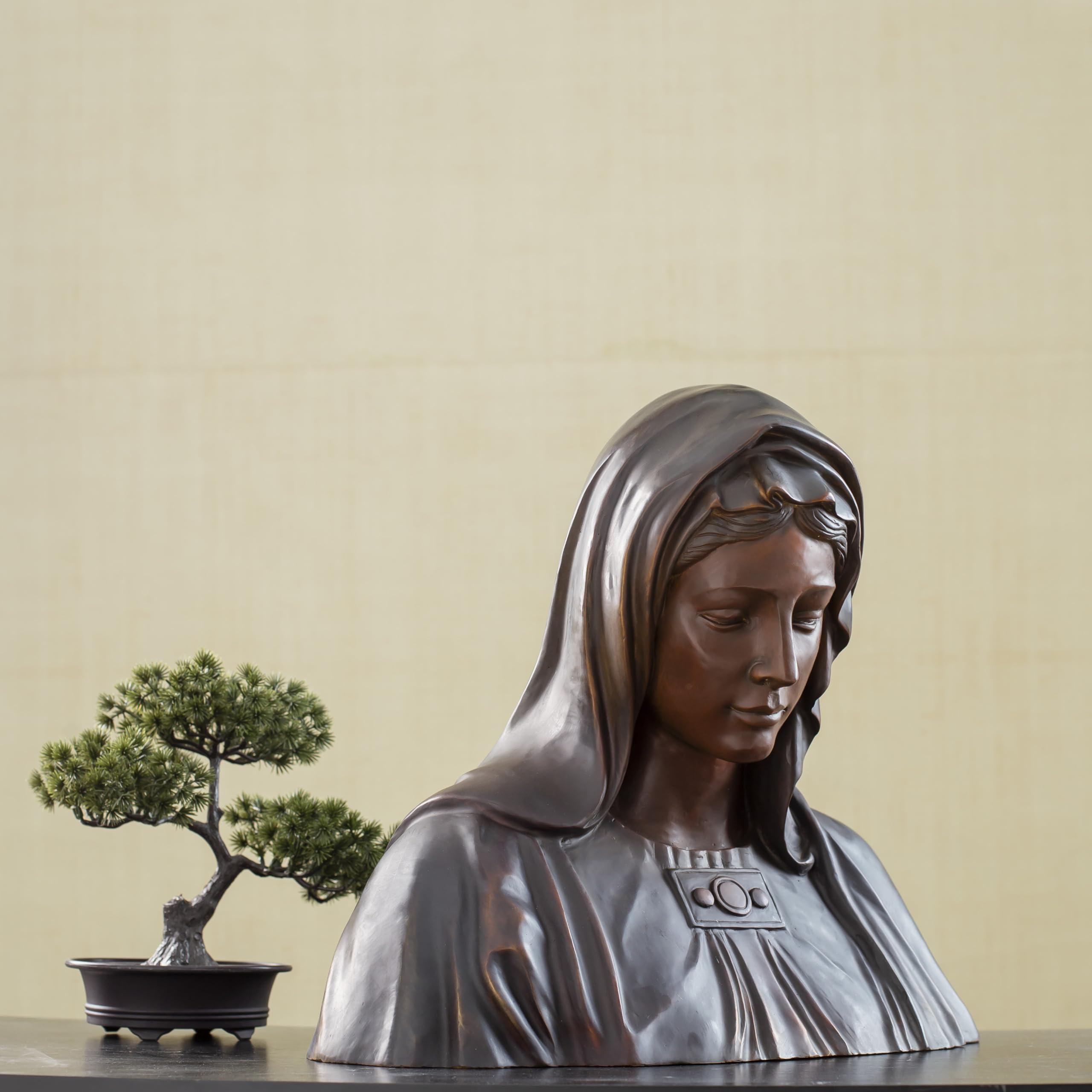 Bronze Virgin Mary bust statue full body view displayed beside bonsai tree in indoor setting