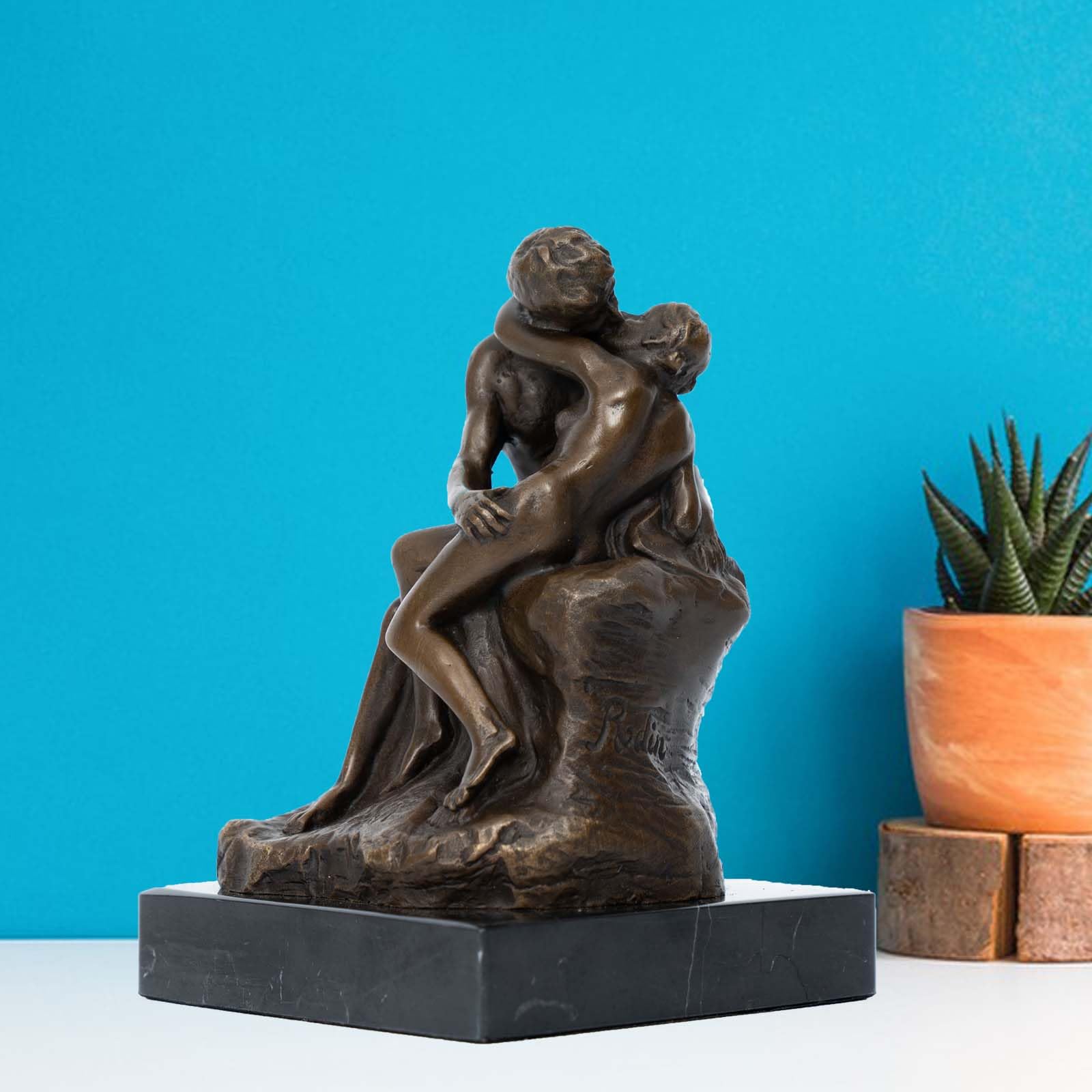 Rodin The Kiss bronze statue on blue background beside potted plant as colorful decor accent