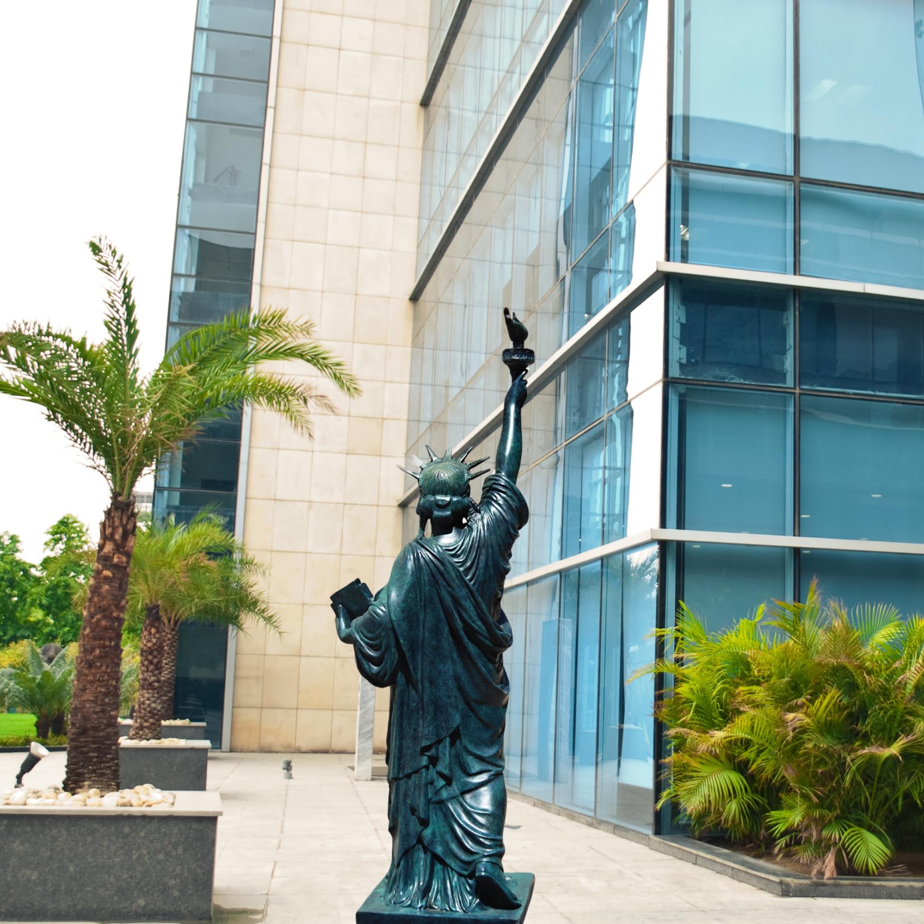 Verdigris blue bronze Statue of Liberty displayed outdoors near modern building with palm trees